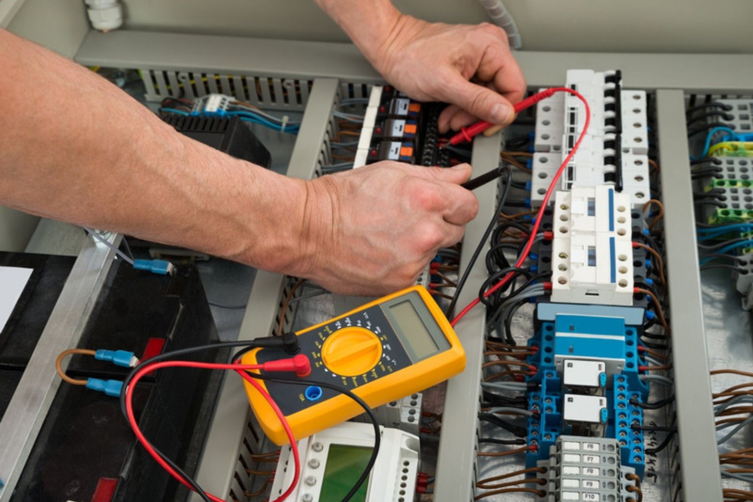 Hænder måler med et gult multimeter på komponenter i en eltavle med mange ledninger