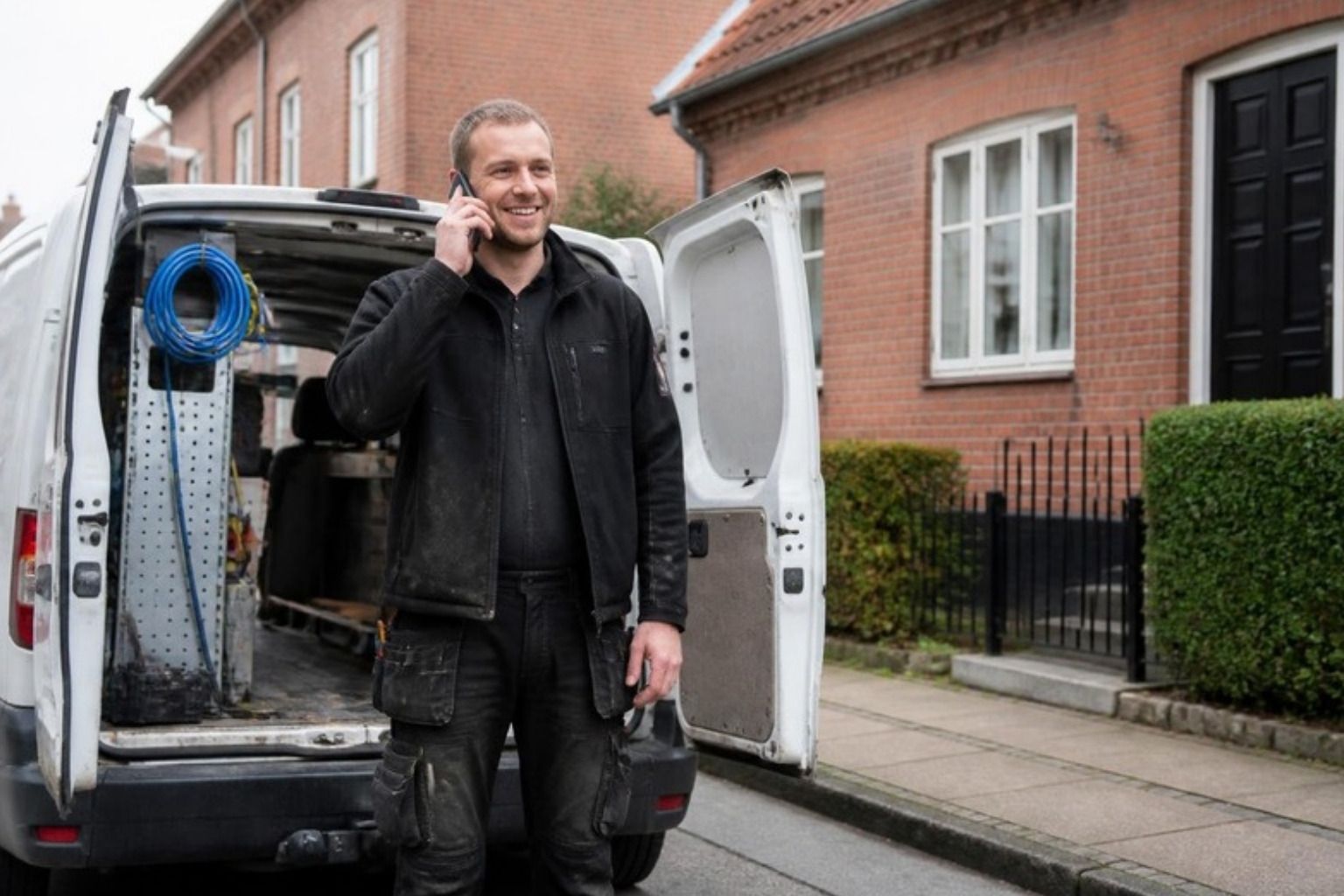 Smilende VVS-installatør taler i telefon ved en åben hvid varevogn foran et rødt murstenshus
